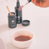 Cosmetic products including a jar, bottle, and brush on a white surface with a bowl of brown powder.